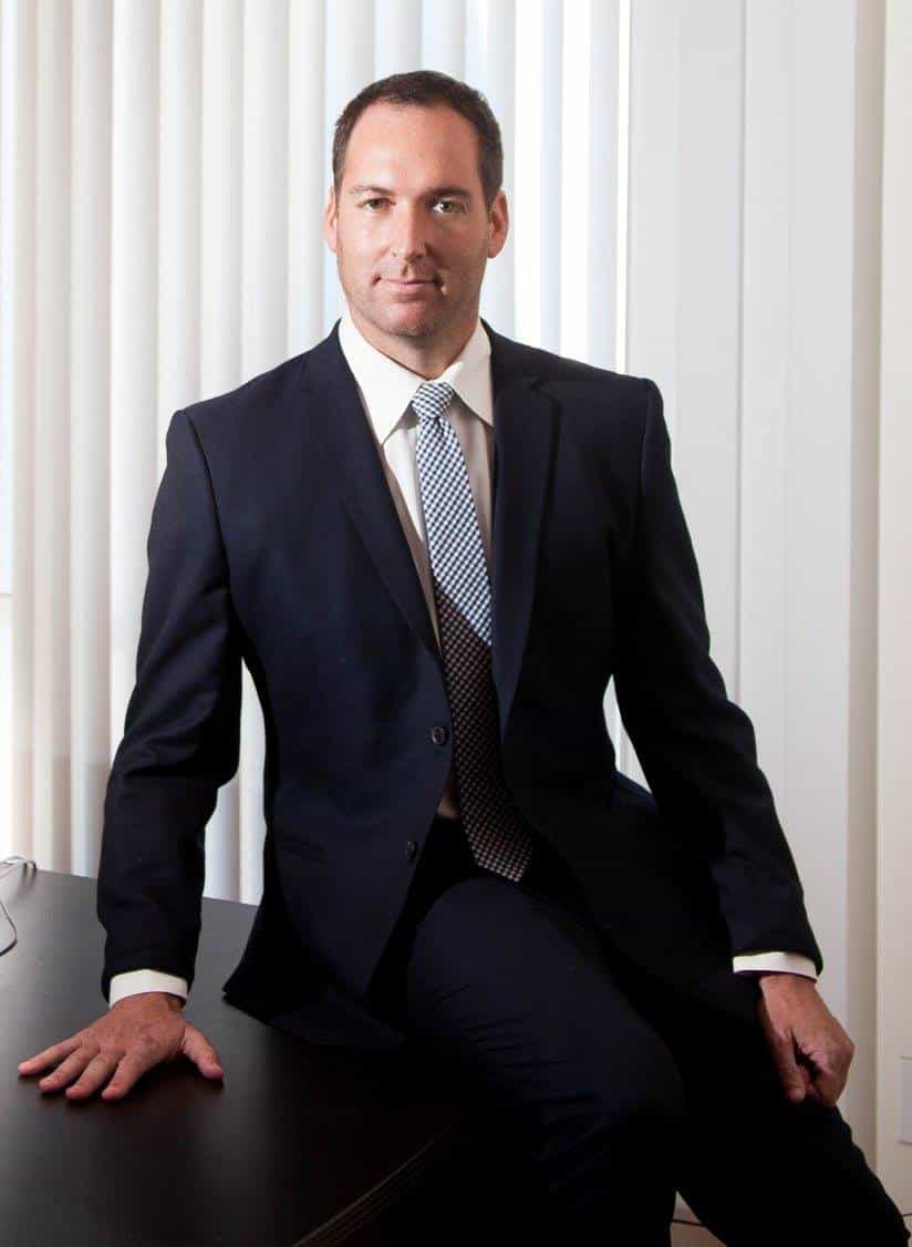 A person in a suit, possibly a Los Angeles employment lawyer, sits on a desk with hands resting. Behind them is a window with vertical blinds, framing the calm yet focused atmosphere of their office.