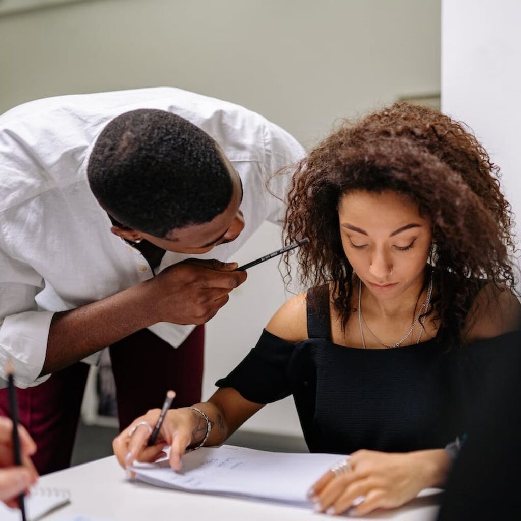 A man leans over to talk to a woman who is writing in a notebook. Both hold pens, clearly engrossed in their discussion. They are seated at a table with papers around them, possibly consulting with an employment lawyer in Los Angeles about workplace rights and regulations. Employment Law